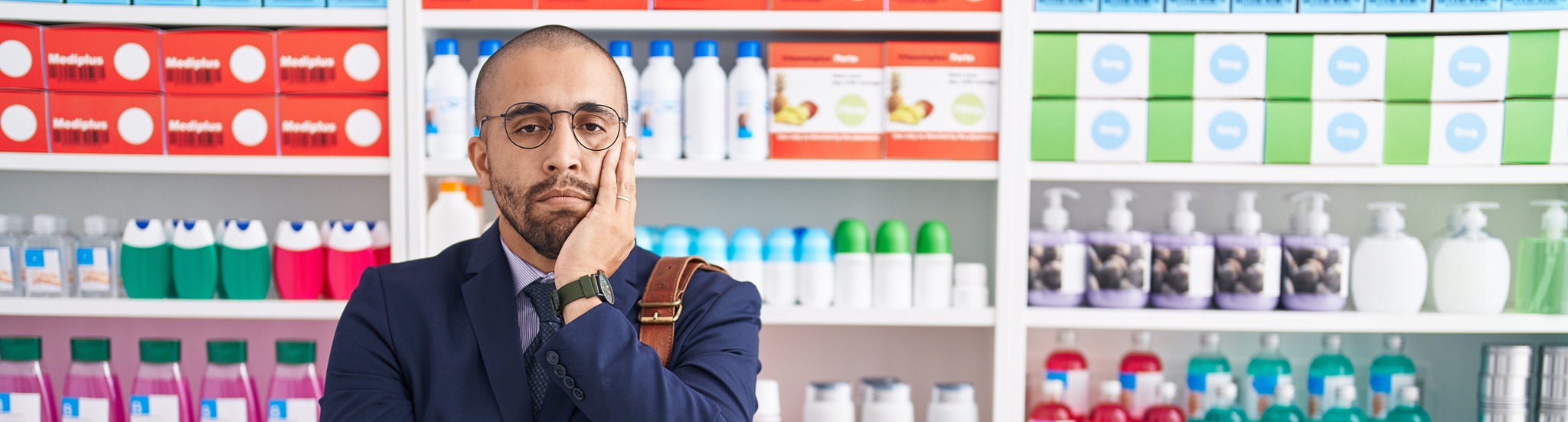 Hispanic Man With Beard Working As Salesman At Pharmacy Drugstore Thinking Looking Tired And Bored With Depression Problems With Crossed Arms.