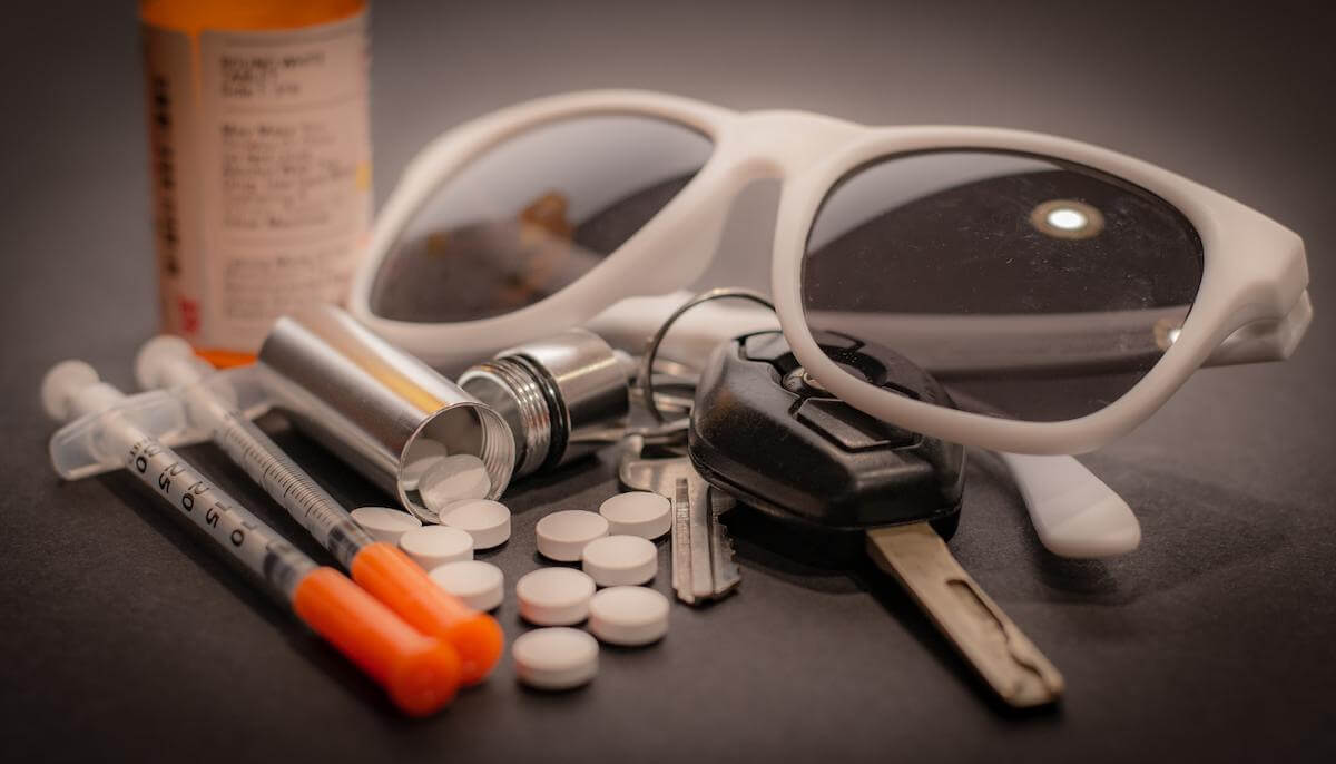 Pills spill out of a container attached to a keyring while syringes and sunglasses lay about.