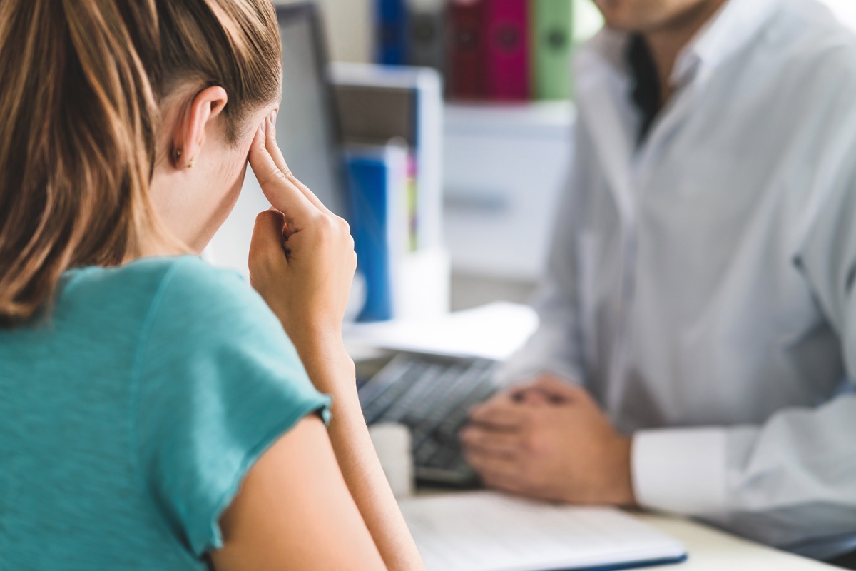 Woman Suffering From Bad Headache Or Migraine. Appointment With Doctor In Office Room. Sick And Unwell Lady With Stress, Trauma Or Burnout. Young Patient In Pain Holding Head With Hands. Medical Visit